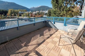 A wooden deck with a blue railing, a white chair, and a view of mountains and buildings in the background. - 1 Bedroom Loft | Fireplace | Village Gate House by Whiski Jack (Whistler)