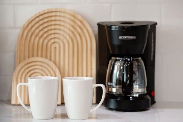 Two white mugs on a countertop with a black coffee maker and a wooden decorative item in the background.