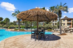 Uma área de piscina com espreguiçadeiras e uma mesa sob um guarda-sol de palha, cercada por palmeiras e um edifício com varandas.