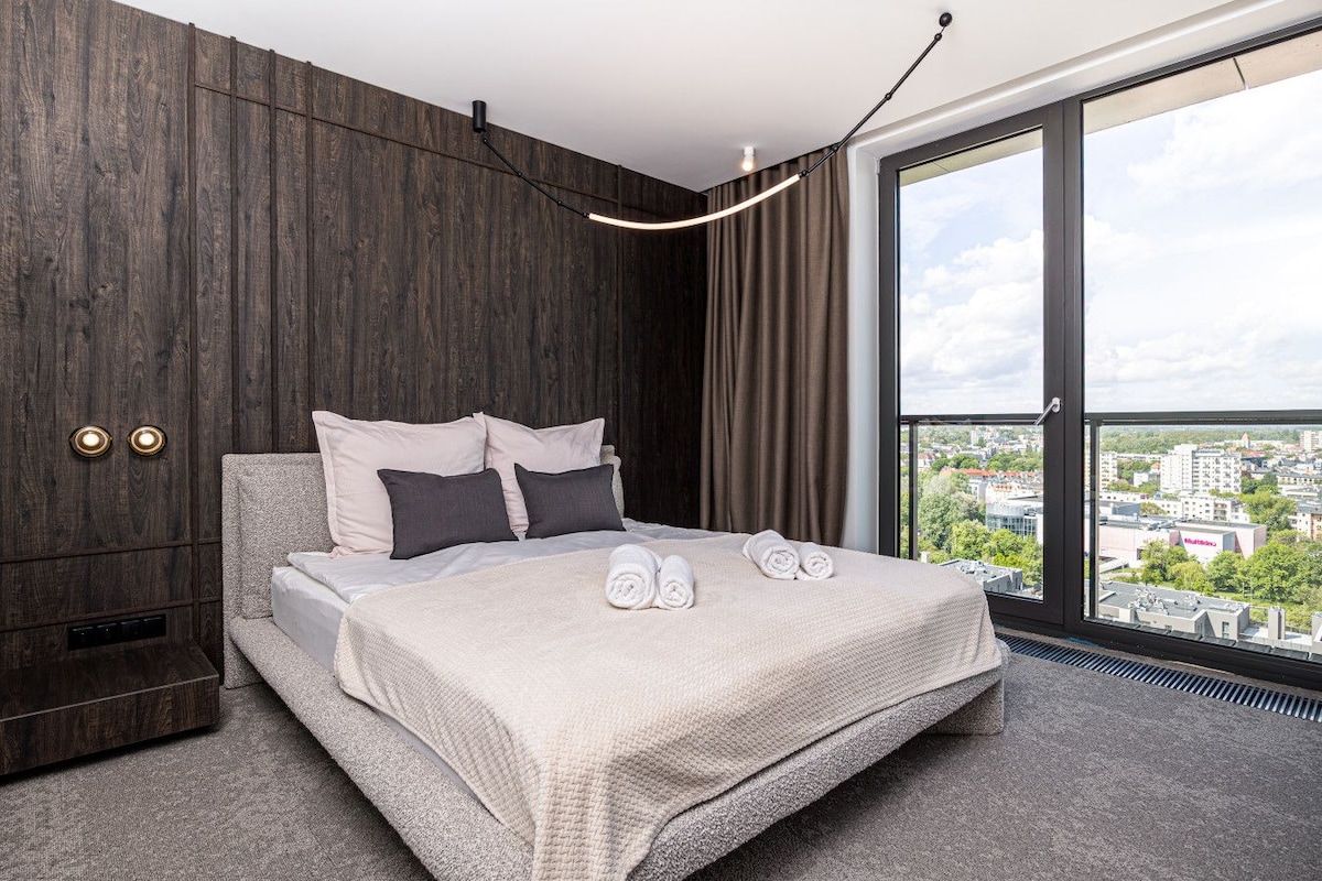 Quarto de hotel moderno com uma cama grande, painéis de madeira escura, vista da cidade e uma janela ampla.