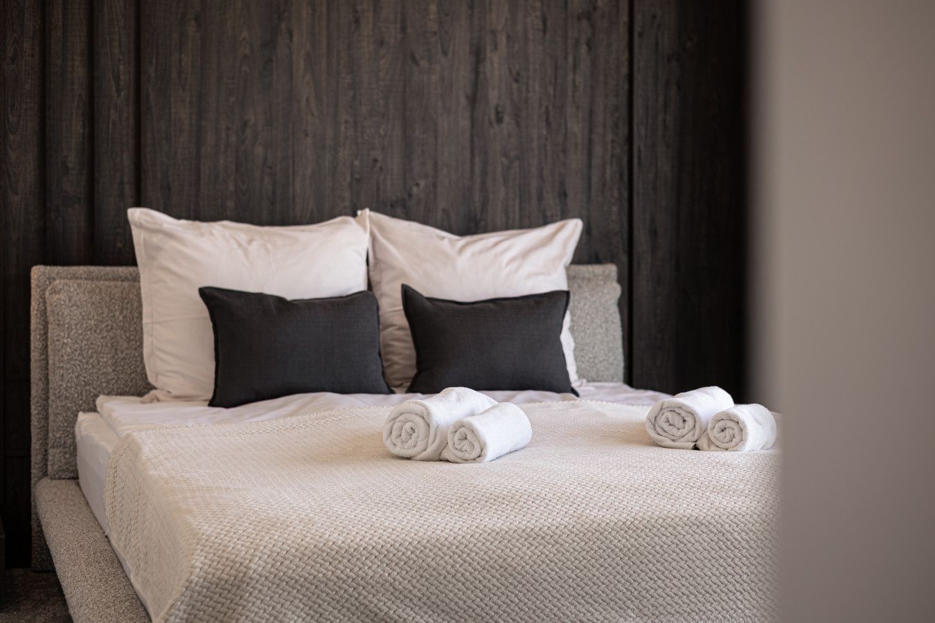 A bed with a textured white cover, two rolled towels, and pillows in white and dark shades.