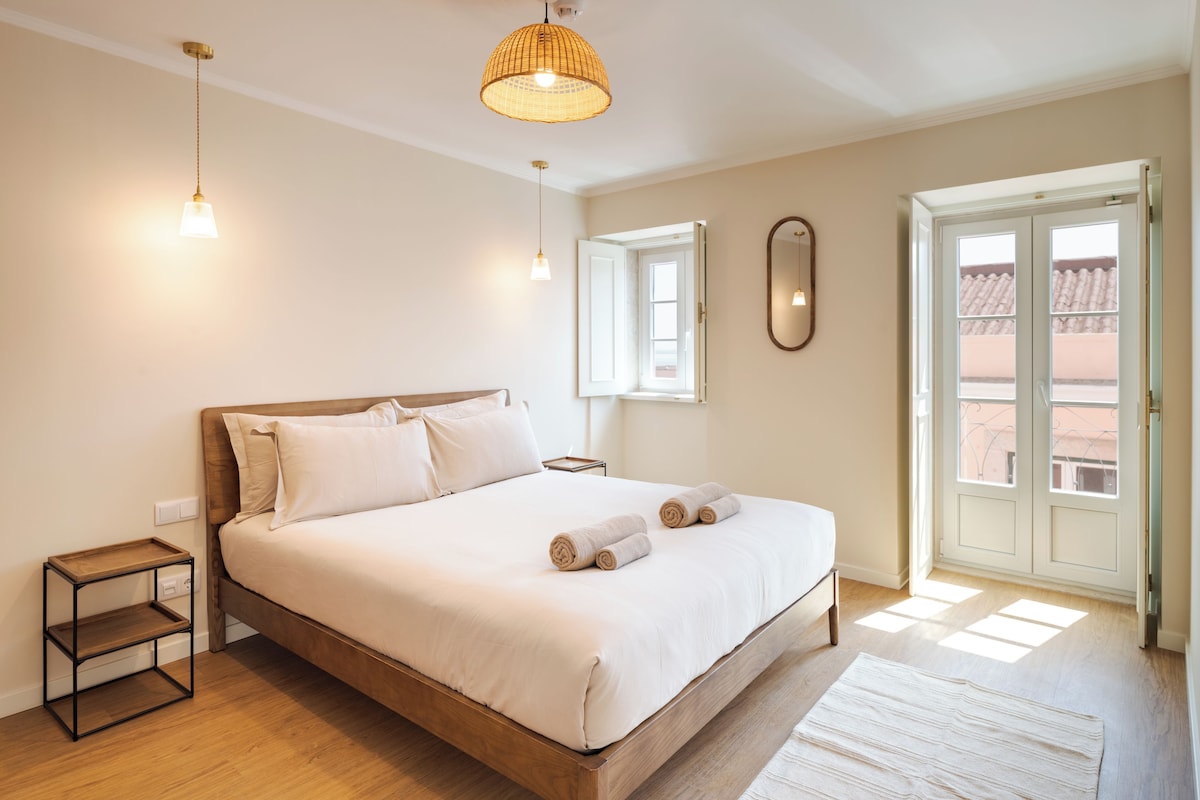 A bedroom with a wooden bed, bedside tables, a hanging light fixture, a window, and a door leading outside.
