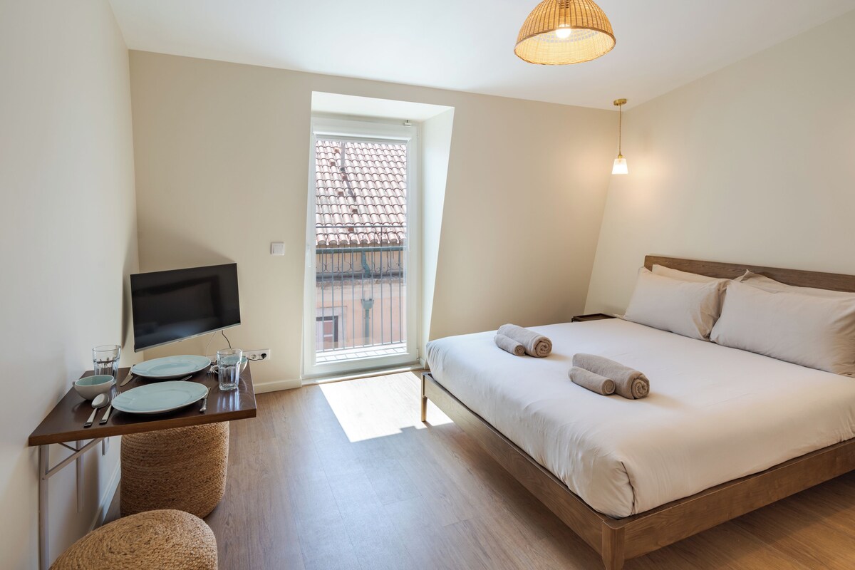 A hotel room with a bed, a television, a dining table with plates and cutlery, and a view of a tiled roof through a window.