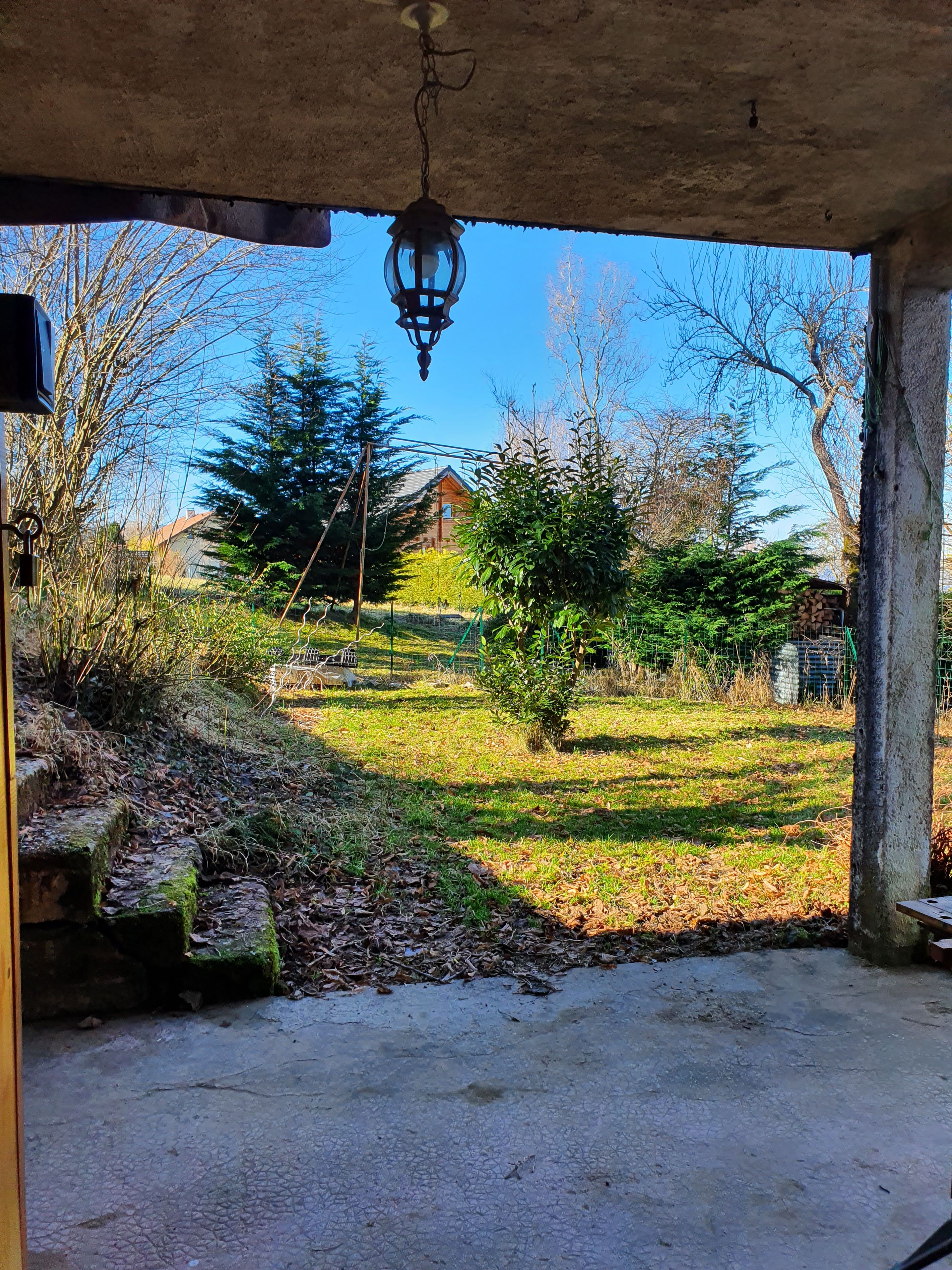 A covered outdoor area with a hanging lantern, a stone bench, and a view of a house and trees.
