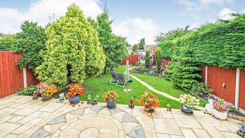 Un patio jardín con suelo de piedra, rodeado por una cerca roja, y diversas macetas con plantas y estatuas de jardín.