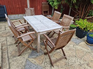 Een houten tafel en stoelen staan op een stenen terras, met een rode hek en bloempotten op de achtergrond.