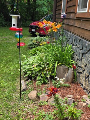 A garden with a bird feeder, a red car parked in the background, and a stone wall. - Private Home,1+acre property,close to WIDells/CastleRock,FirePit,yard games,wood (Adams)