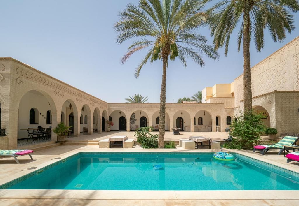 A pool area with a clear blue pool, palm trees, and a building with arches.