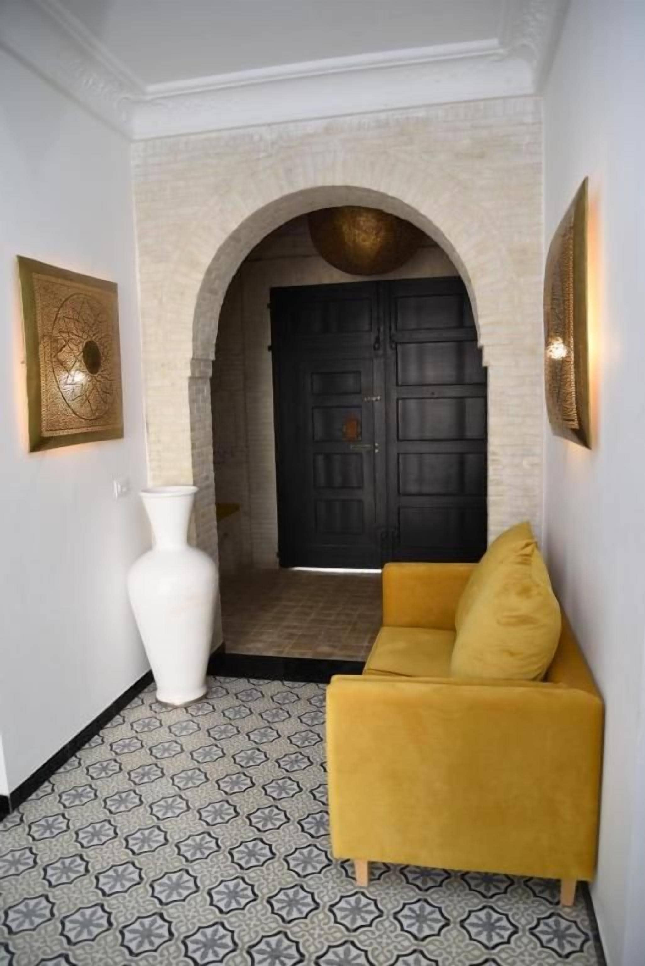 A hallway with a white vase, a yellow sofa, and a black door with a decorative archway.