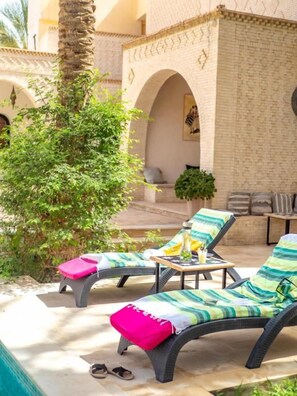 A sunlit outdoor lounge area with two lounge chairs, a small table, and a potted plant.