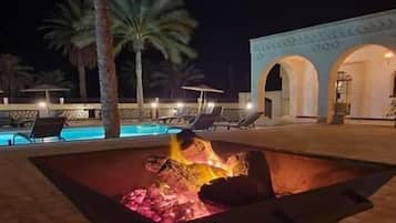A nighttime outdoor area with a fire feature, palm trees, and a pool.