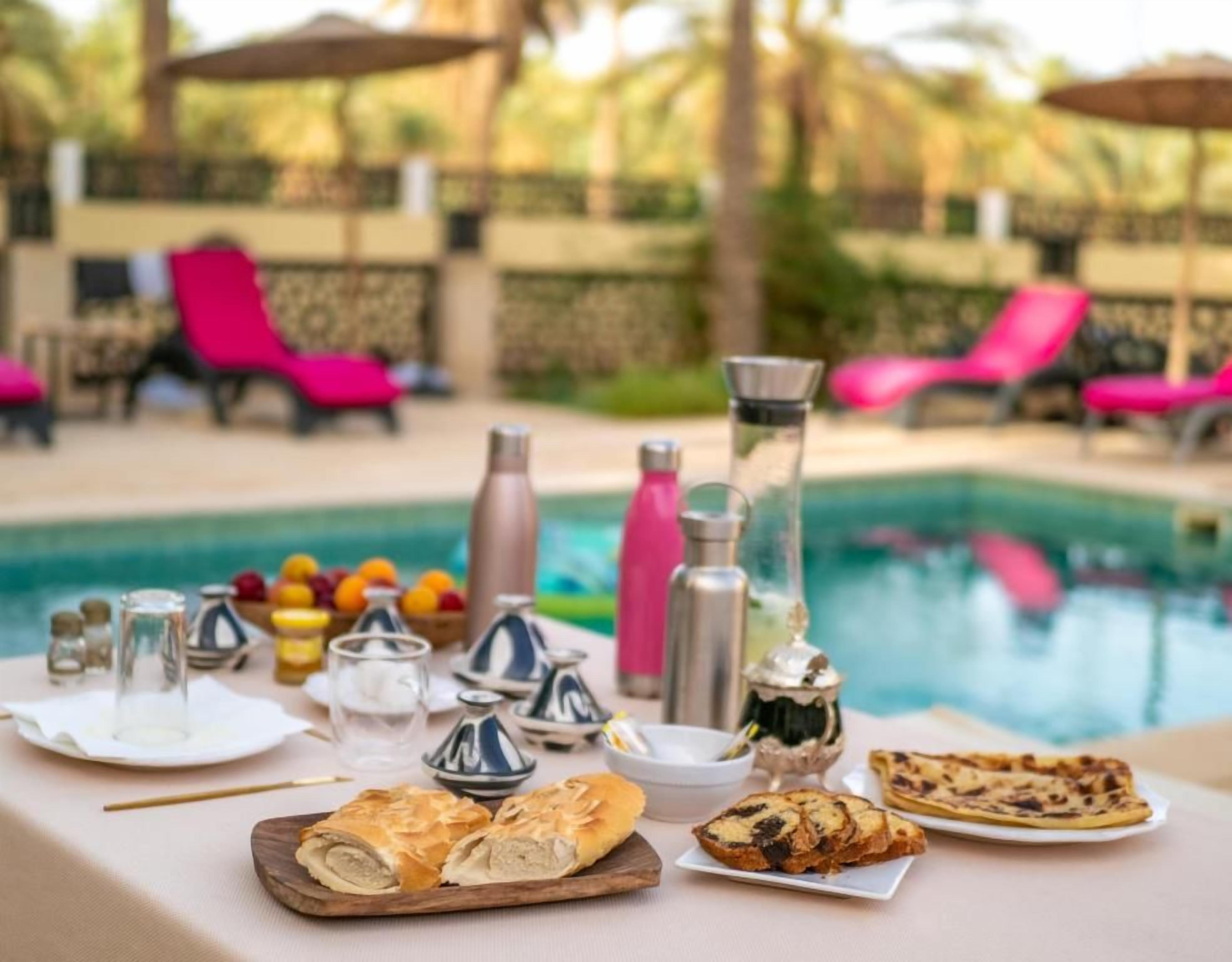 A table set with food and drinks by a poolside, with lounge chairs and a pool in the background.