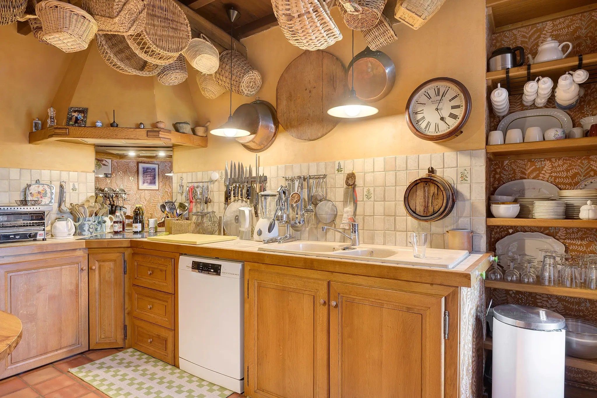 Een keuken met houten kastjes, een vaatwasser, hangend keukengerei en een klok aan de muur.