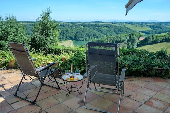 Two outdoor chairs with a table and bottles, overlooking a scenic landscape with hills and a lake.