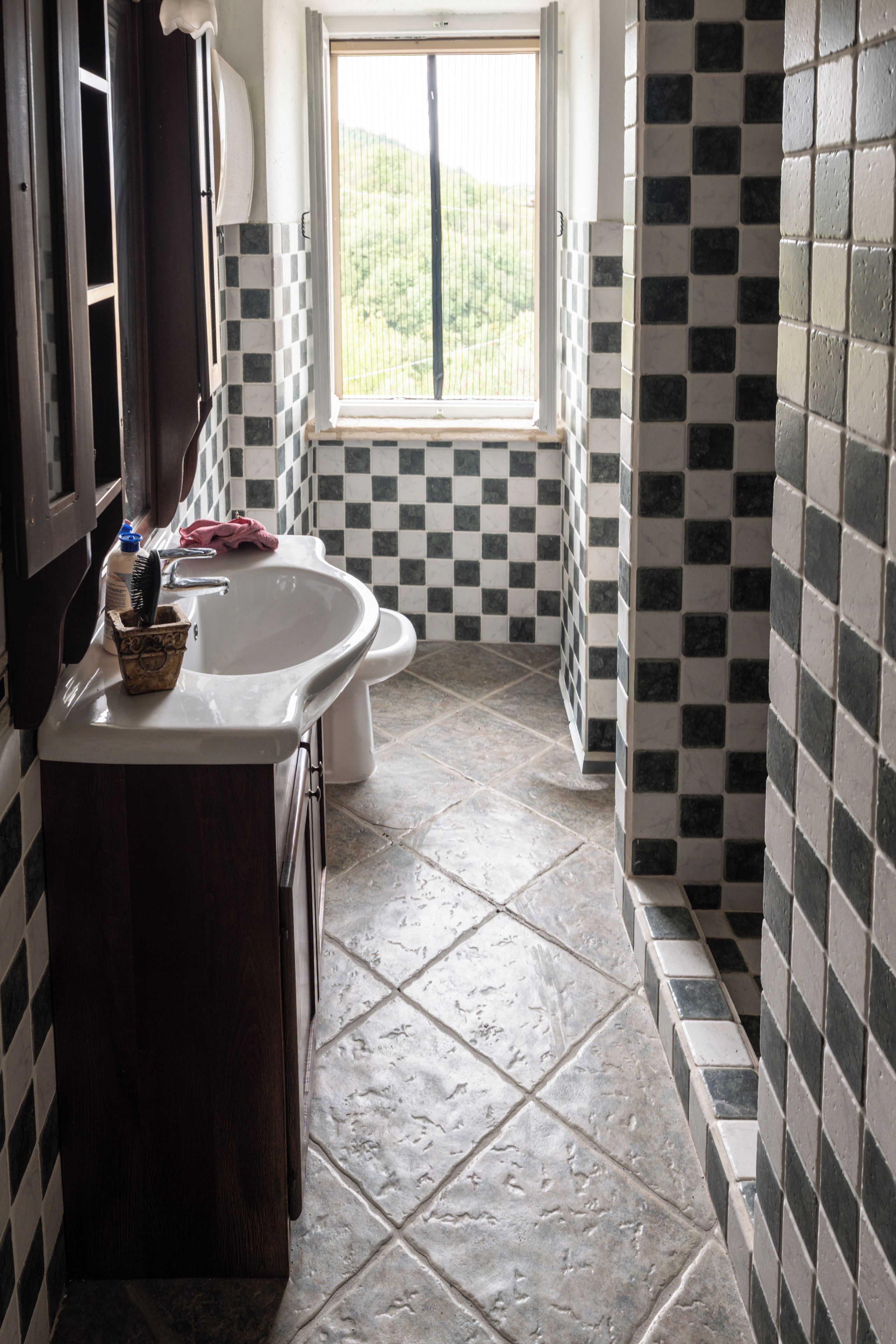 Un baño con piso a cuadros, un lavamanos y una ventana con vista a la vegetación.