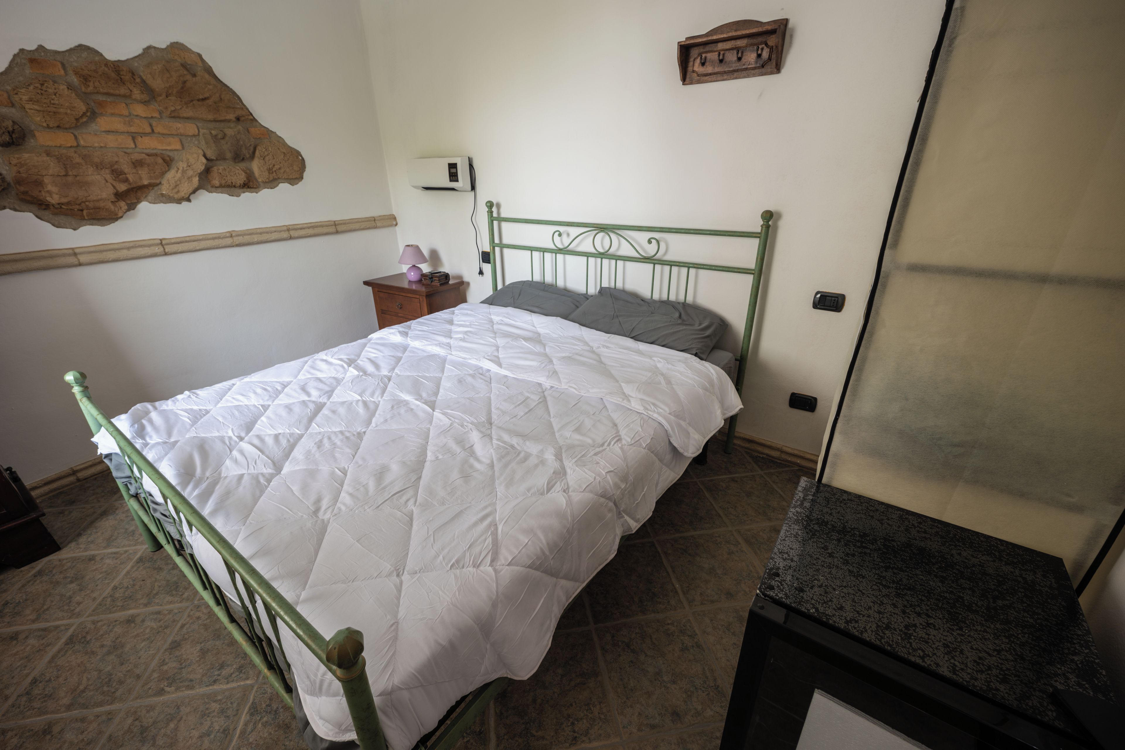 A bedroom with a bed, a nightstand, a wall-mounted heater, and a mirror.