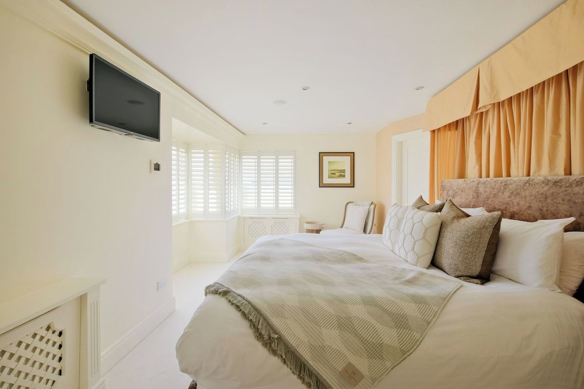 A bedroom with a large bed, a television mounted on the wall, and a window with blinds.