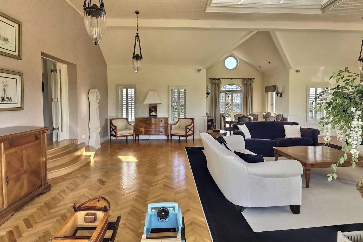 A spacious living room with a wooden herringbone floor, a dark blue sofa, and a white armchair. There is a wooden cabinet and a small table with a lamp.