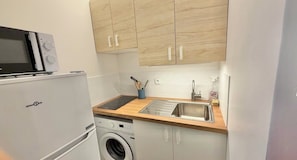 A compact kitchen with a microwave, washing machine, and wooden cabinets. - Renovated apartment with a terrace and parking (Houlgate)