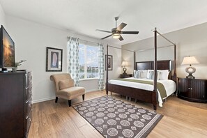 A four-poster bed with a dark headboard, a ceiling fan, a view of the outdoors through a window with patterned curtains, and a dark wooden dresser with a flat-screen TV. - Amazing 4BR at Vista Cay with Gorgeous Lake View (Orlando)