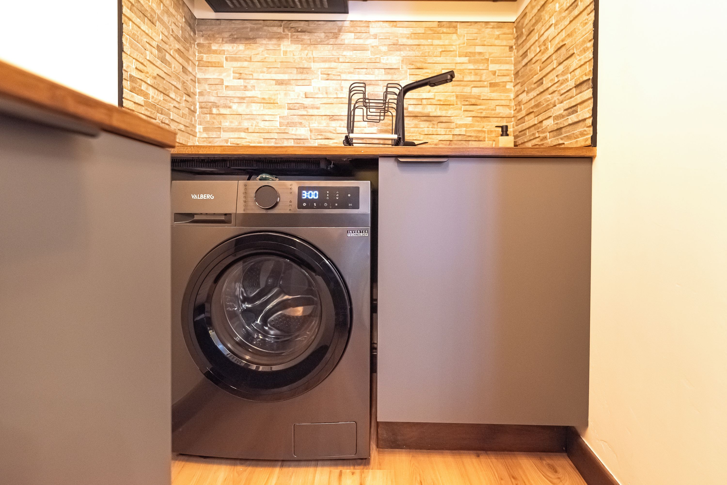 Une petite buanderie équipée d’un lave-linge à chargement frontal, d’un petit meuble de rangement et d’un pare-feu en briques.