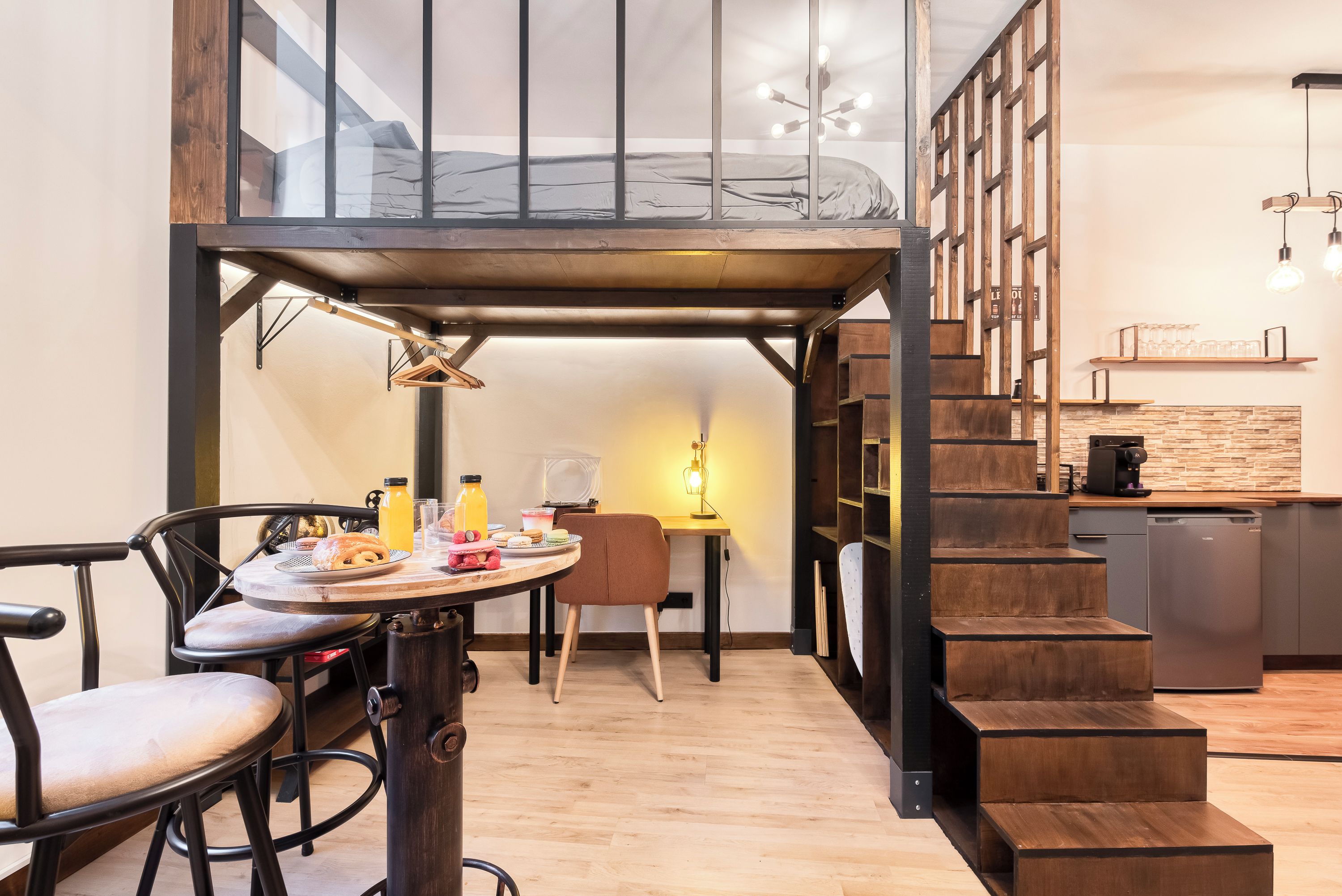 A compact living space with a loft bed, a dining area with a table and chairs, and a kitchen area with a dishwasher and a coffee machine.