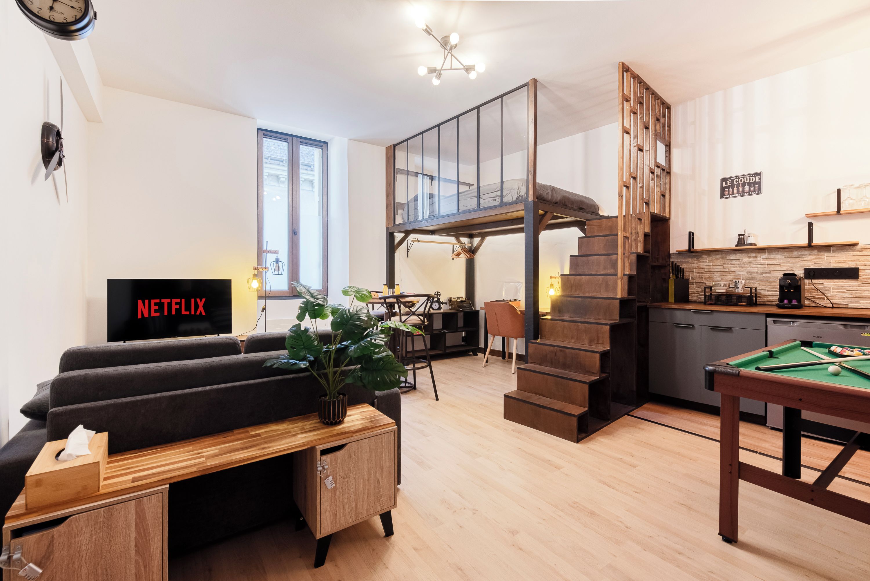 A modern living space with a sofa, a coffee table, a television, a staircase, a kitchen area, and a pool table.