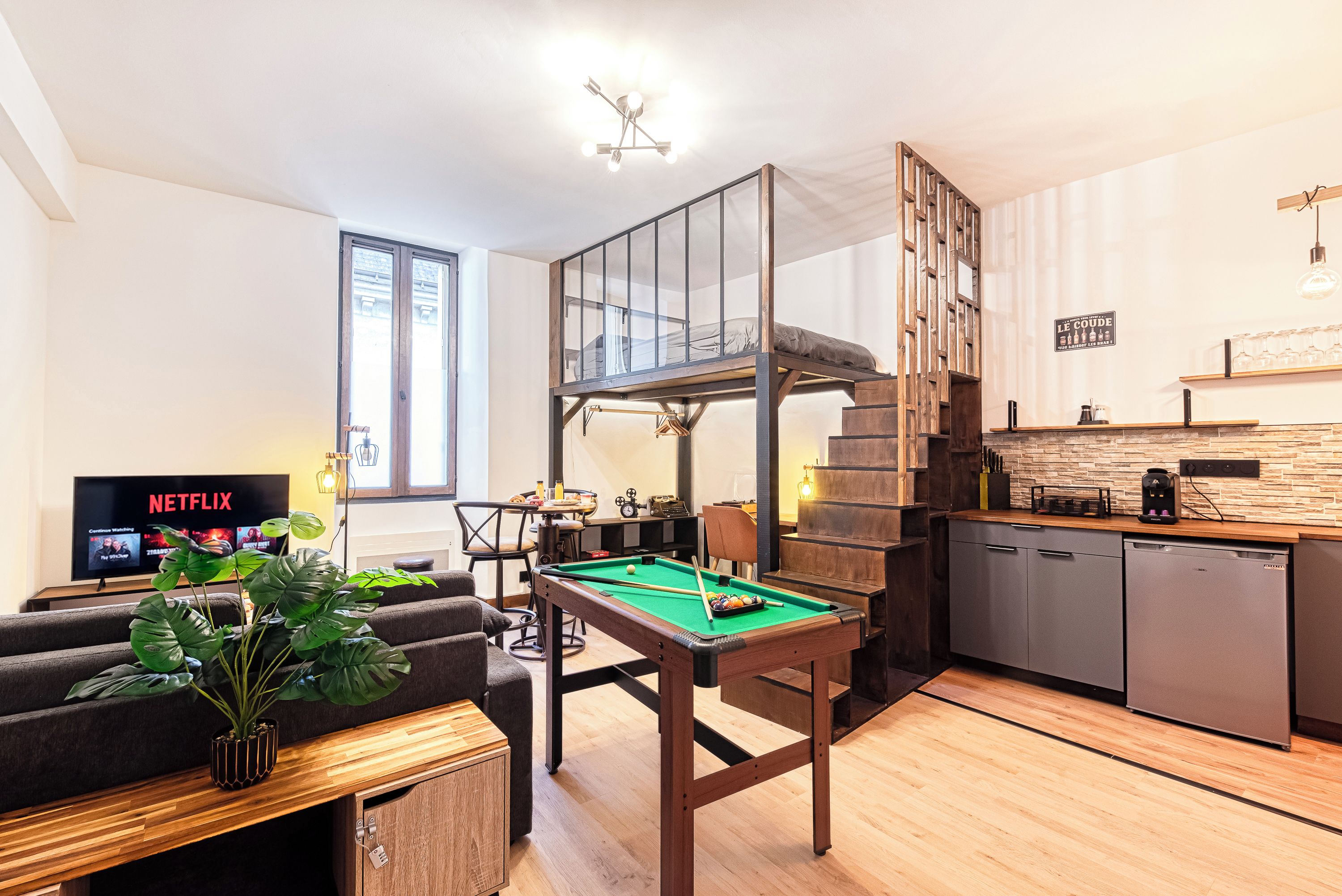 Ein modernes Wohnzimmer mit Billardtisch, einer Couch, einem Fernseher, der eine Netflix-Sendung zeigt, und einer Treppe, die zu einer oberen Ebene führt.