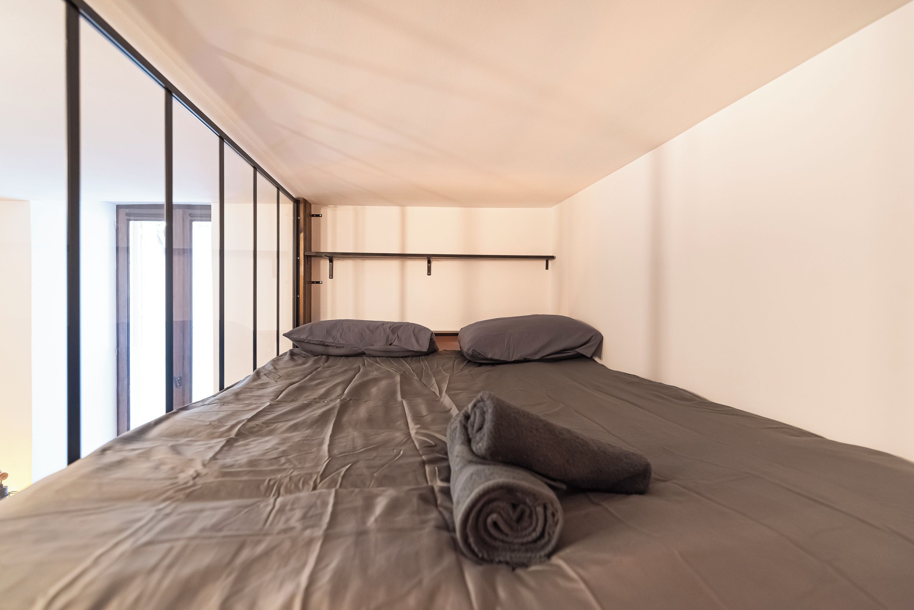 Een eenpersoonsbed met een grijze matras en kussens, een opgerolde grijze deken, en een hoofdbord met een horizontale lamp.