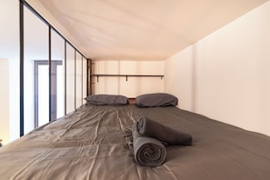 Ein Einzelbett mit grauem Matratze und Kissen, eine zusammenrollte graue Decke und ein Kopfteil mit einer horizontalen Leuchte.