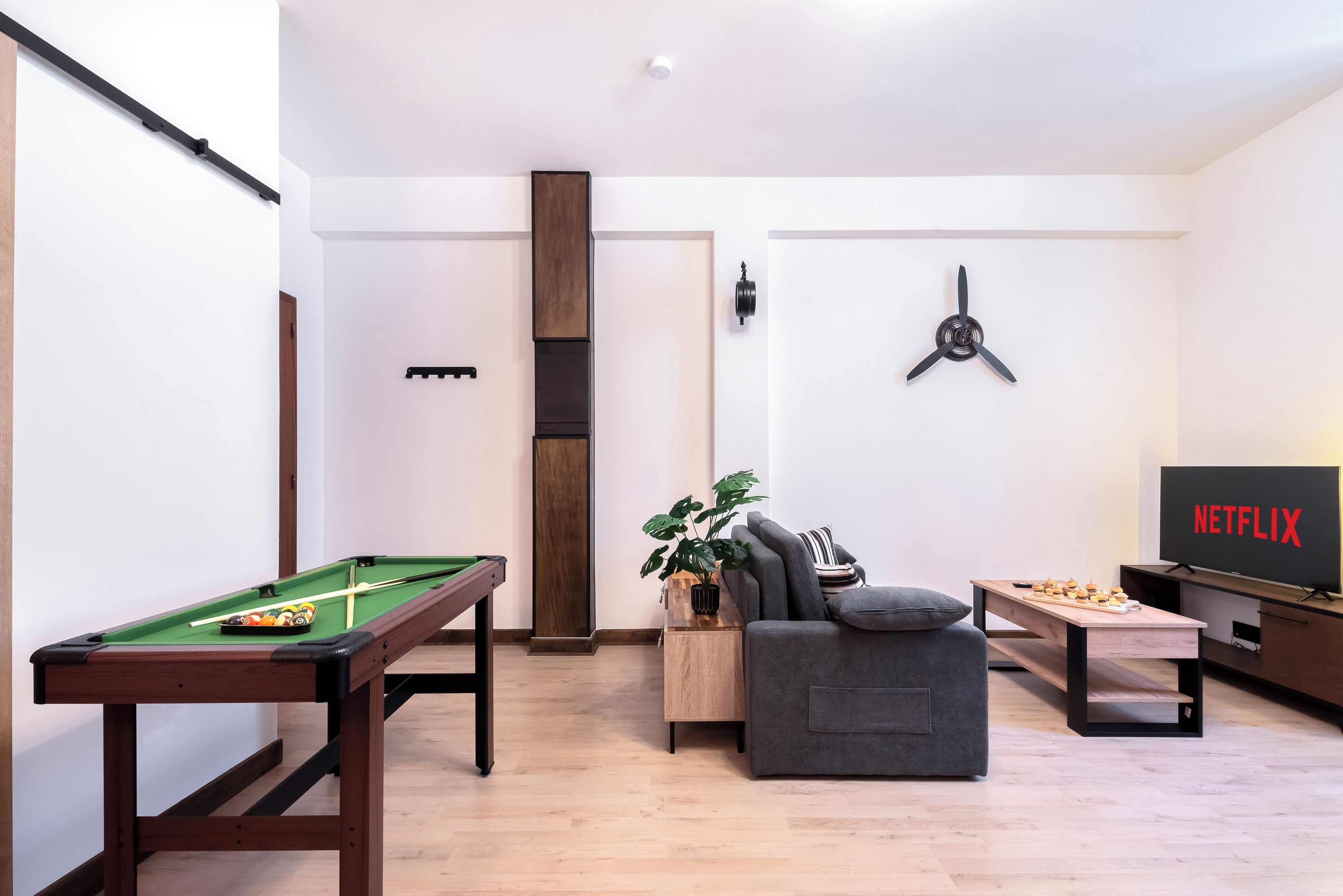 A modern living room with a pool table, a sofa, a coffee table, a plant, and a TV displaying the Netflix logo.