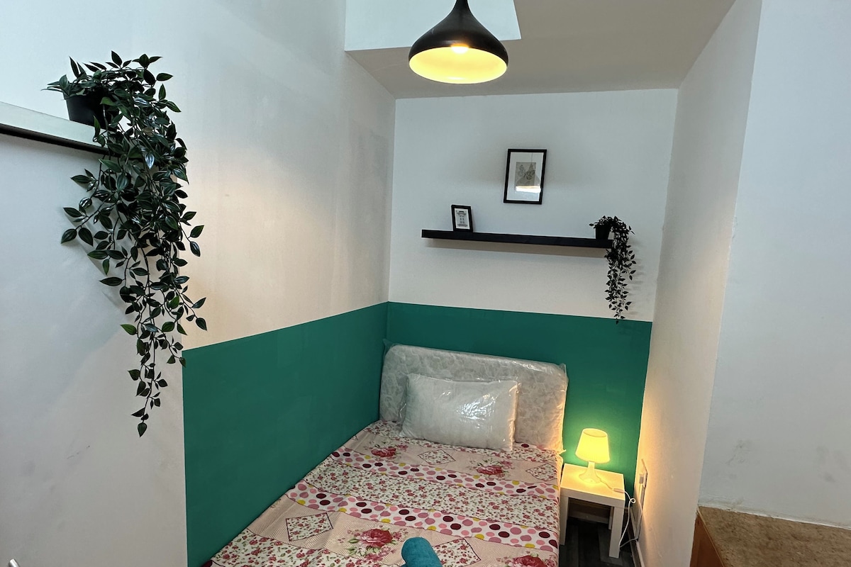 A small, cozy room with a bed, a nightstand, a wall-mounted shelf, and a hanging plant.