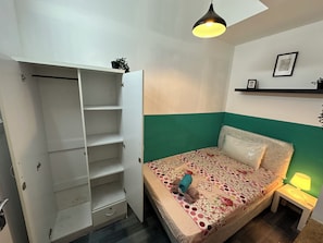 A bedroom with a bed, a nightstand, a lamp, a shelf with a picture, and a wardrobe. - Bagpack Partition Room in Tecom (Dubai)