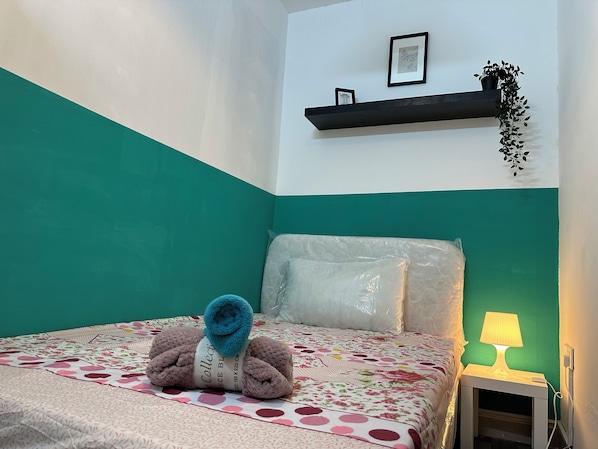Una cama con colcha de estampado floral, una toalla enrollada y una almohada. Hay una mesita de noche con una lámpara y un enchufe.