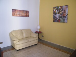 Ein Zimmer mit einem beigen Sofa, einem kleinen Tisch mit einer Lampe und zwei an der Wand hängenden Kunstwerken.