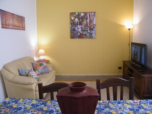 A living room with a sofa, a table with a patterned tablecloth, a television on a wooden stand, and a painting on the wall. - Holiday Home 'Mi manda Picone da Lolli's Liberty Apartment' with Balcony and Air Conditioning (Palermo)