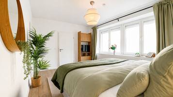 Ein Schlafzimmer mit Bett, Fernseher, Fenster mit Vorhängen und einer Topfpflanze.