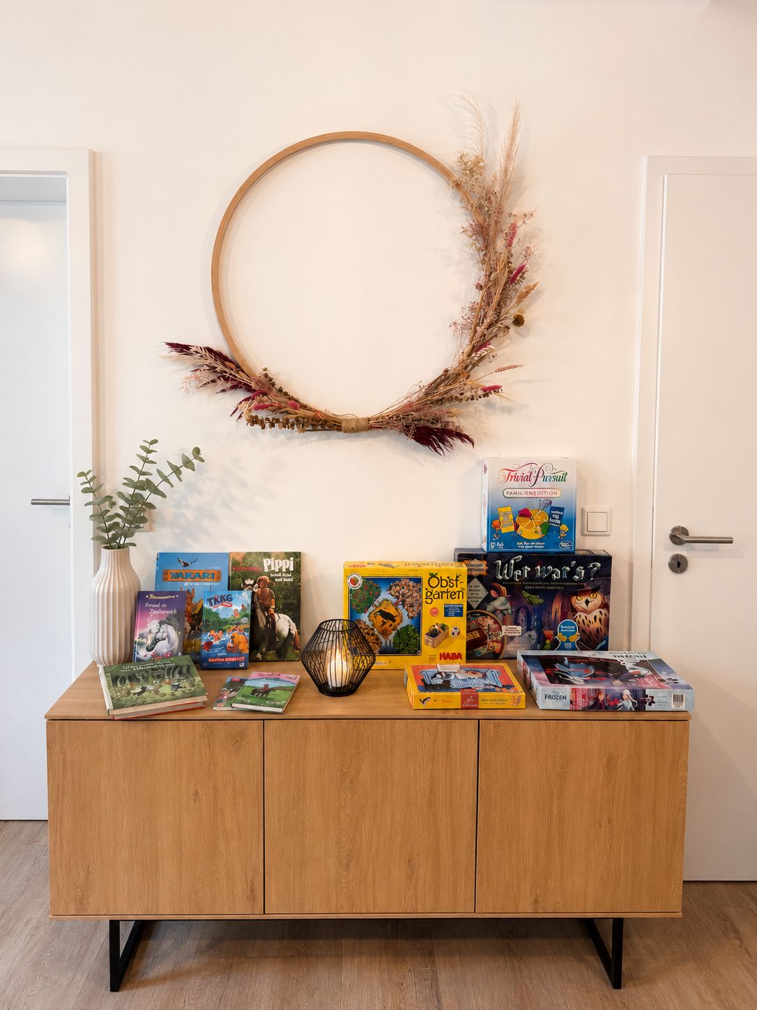 Ein Holzkabinett mit verschiedenen Gesellschaftsspielen und Büchern, ein dekorativer Kranz an der Wand und eine Vase mit getrockneten Pflanzen.