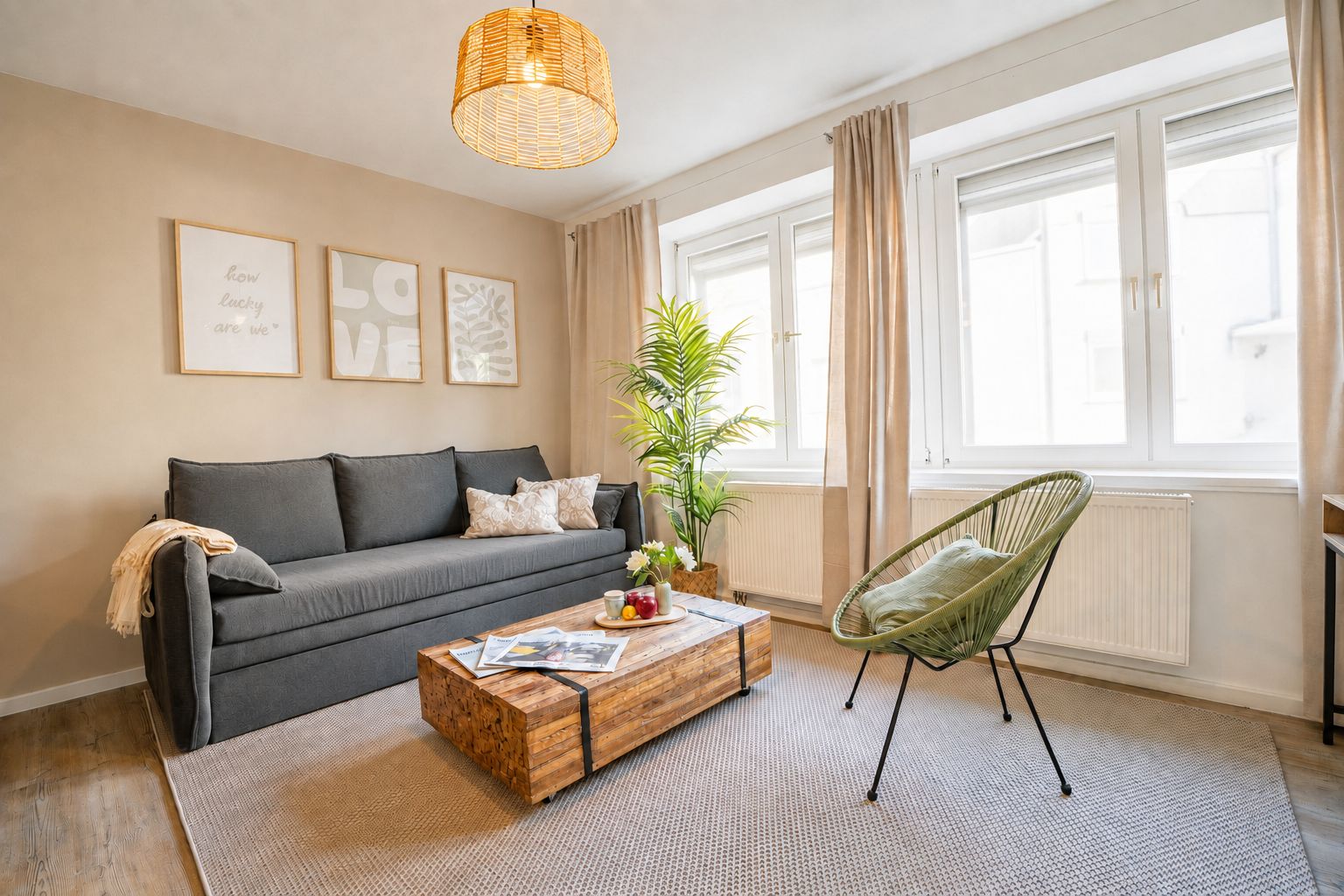 Ein Wohnzimmer mit einer Couch, einem Holztisch, einem grünen Sessel, einer Pendelleuchte und einem Fenster mit Vorhängen.