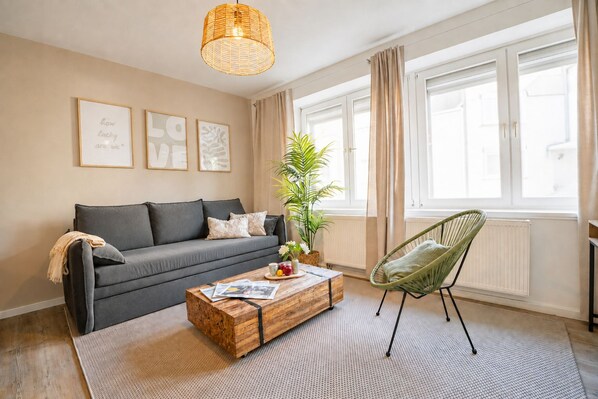 A living room with a sofa, a wooden coffee table, a green chair, a hanging light fixture, and a window with curtains.
