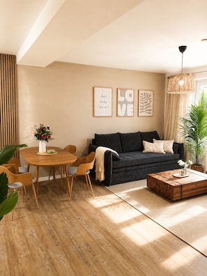 Un salon moderne avec un canapé foncé, une table à manger en bois et des chaises. On peut y voir des tableaux encadrés au mur, une plante en pot et un luminaire suspendu.