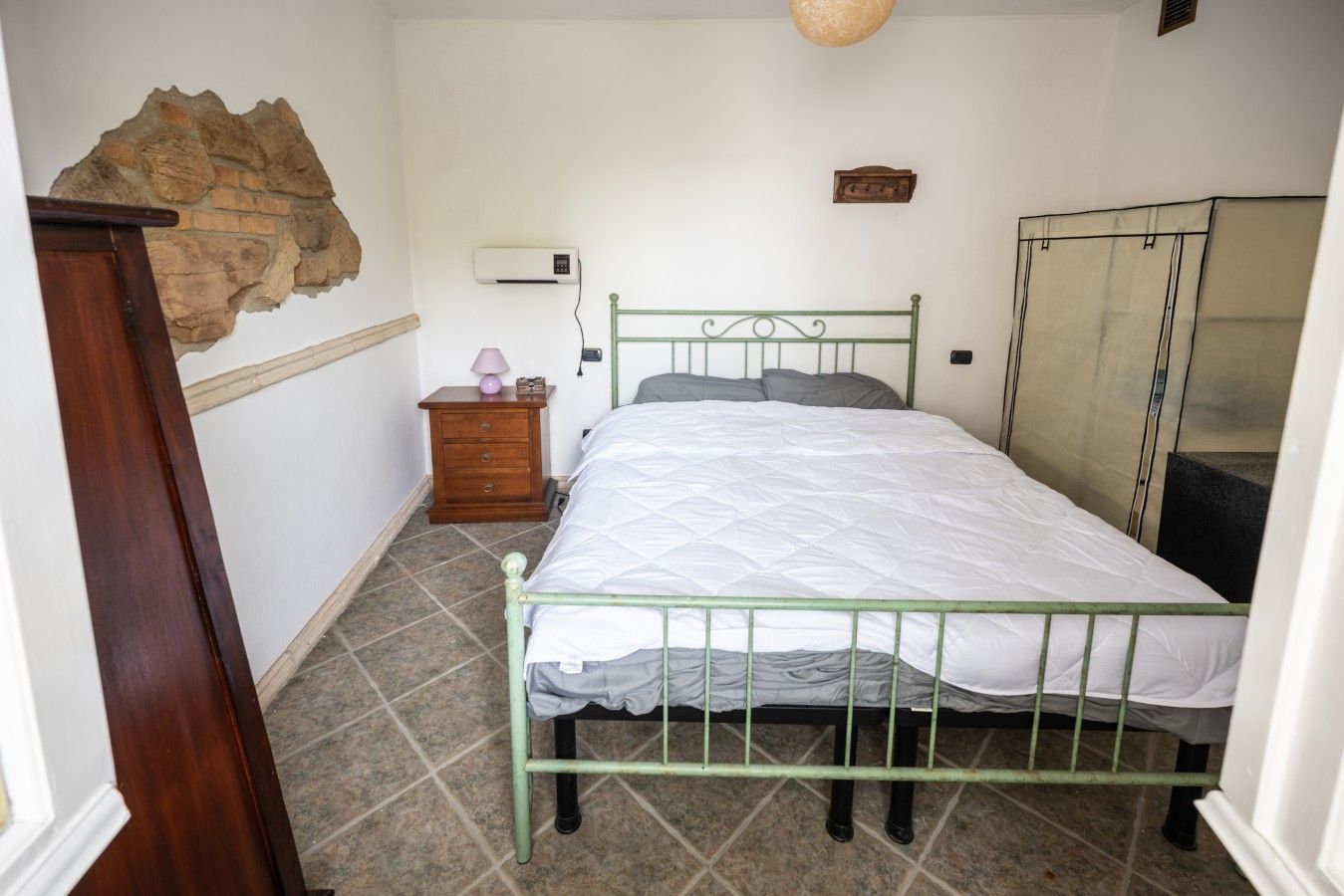 Ein Schlafzimmer mit einem grünem Metallbettgestell, einem Nachttisch aus Holz mit einer Lampe und einer Steinwand.