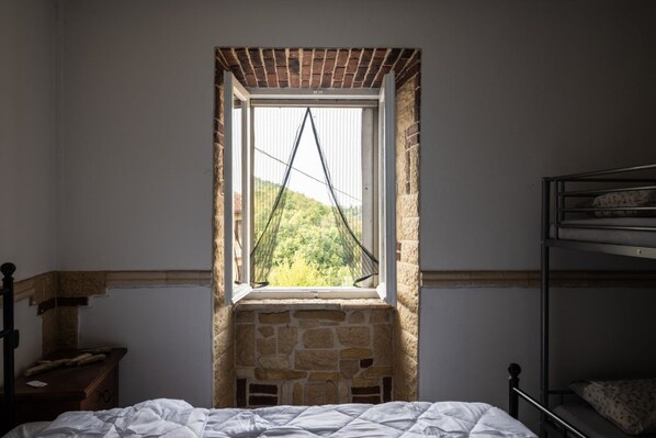 A bedroom with a stone wall, a window with a view of greenery, a bunk bed, and a wooden nightstand.