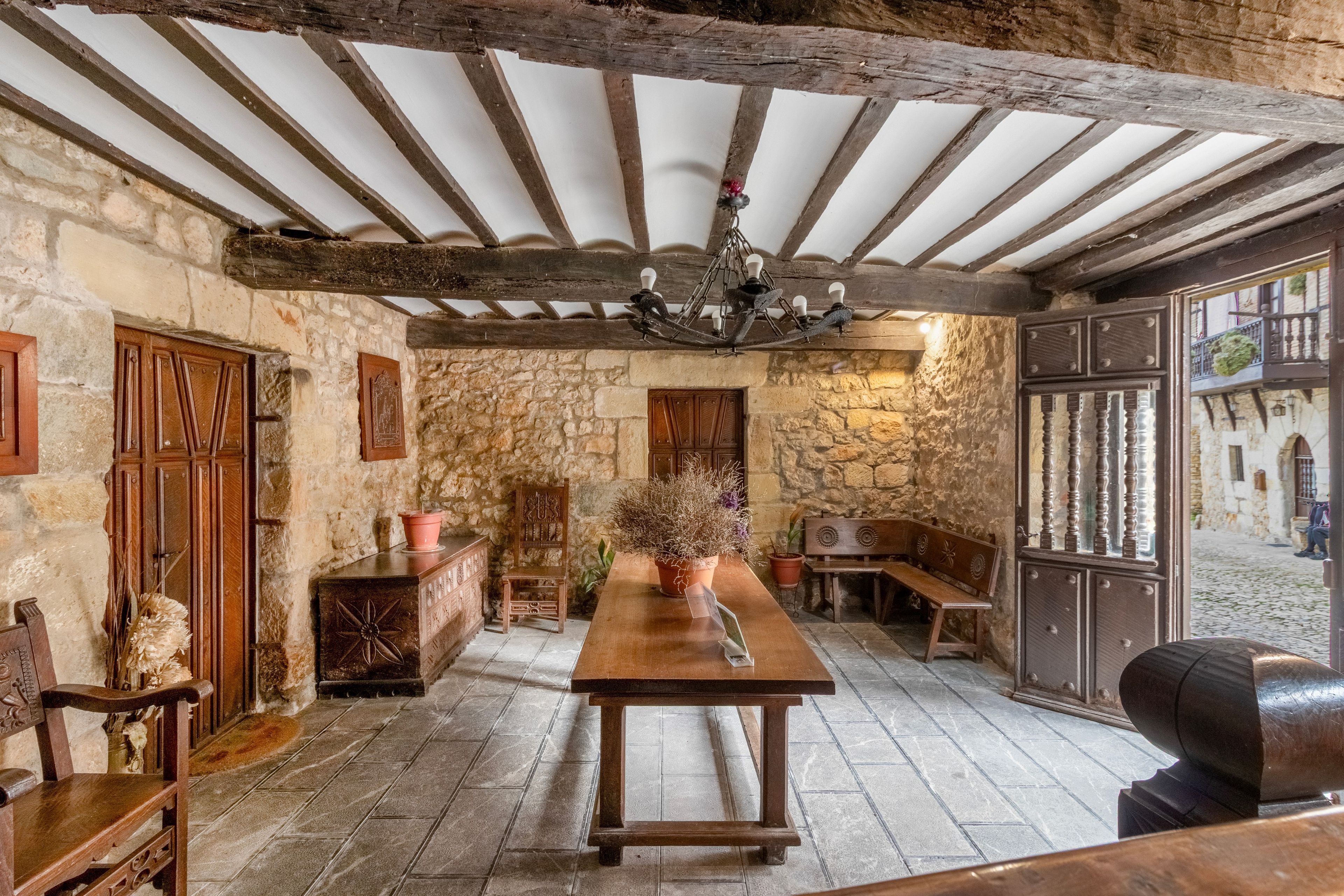 Un interior rústico con paredes de piedra vista, vigas de madera, una mesa de madera, bancos y una araña.