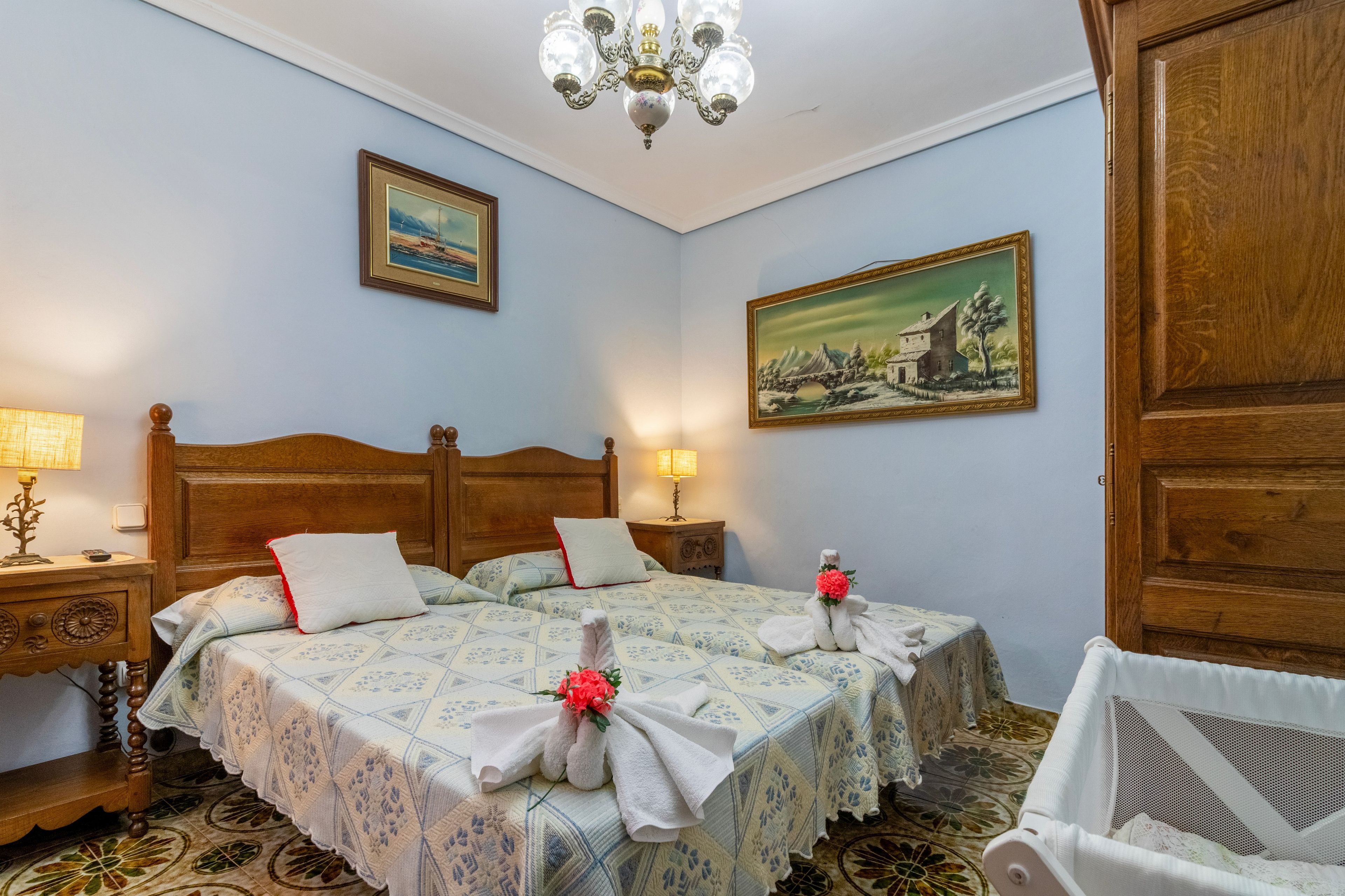 A hotel room with a wooden bed, two bedside tables, a chandelier, and two paintings on the walls.