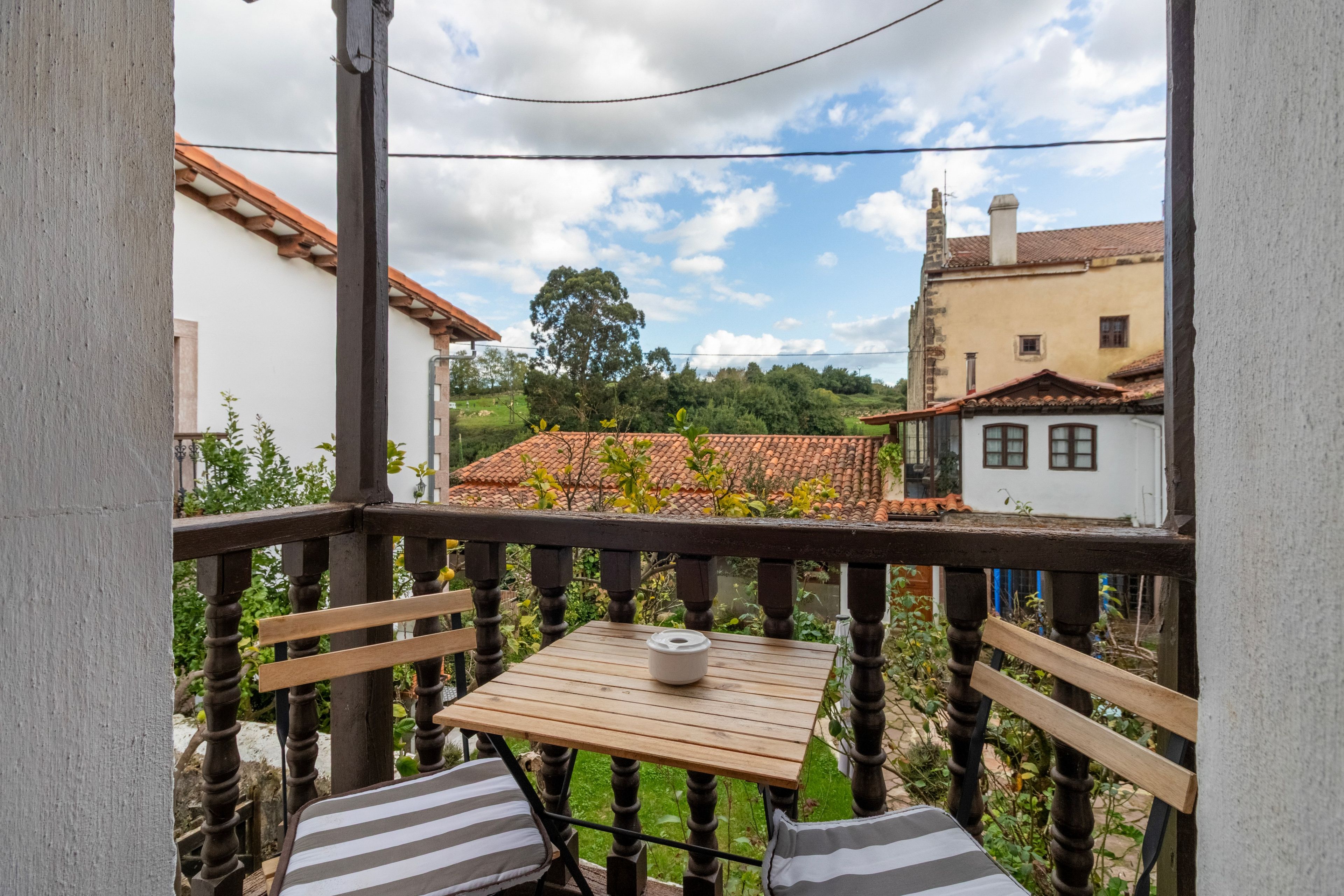 Un balcon avec une table en bois et deux coussins à rayures, donnant sur un quartier résidentiel avec des toits en tuiles et de la verdure.