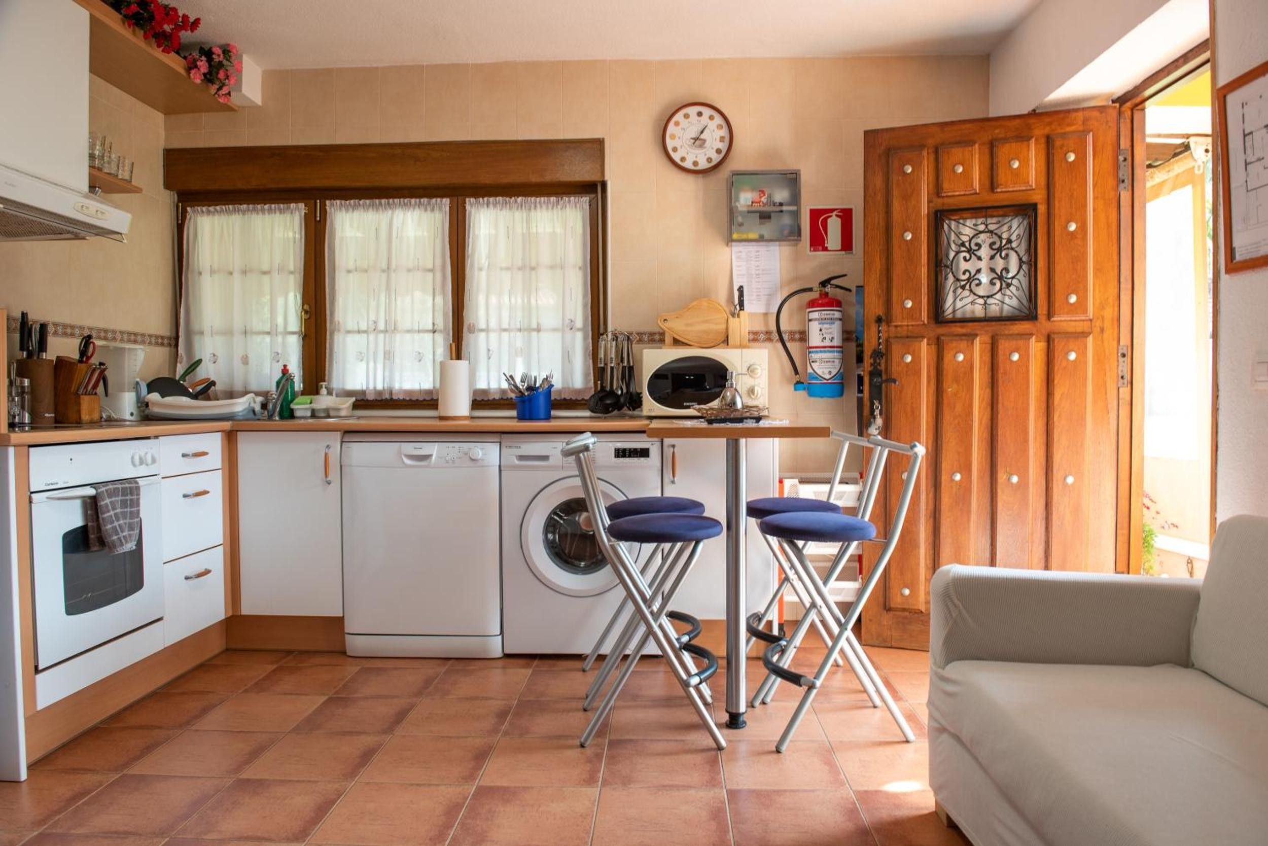 Cocina compacta con lavadora, microondas y lavaplatos. Hay dos taburetes de bar y un sofá blanco.