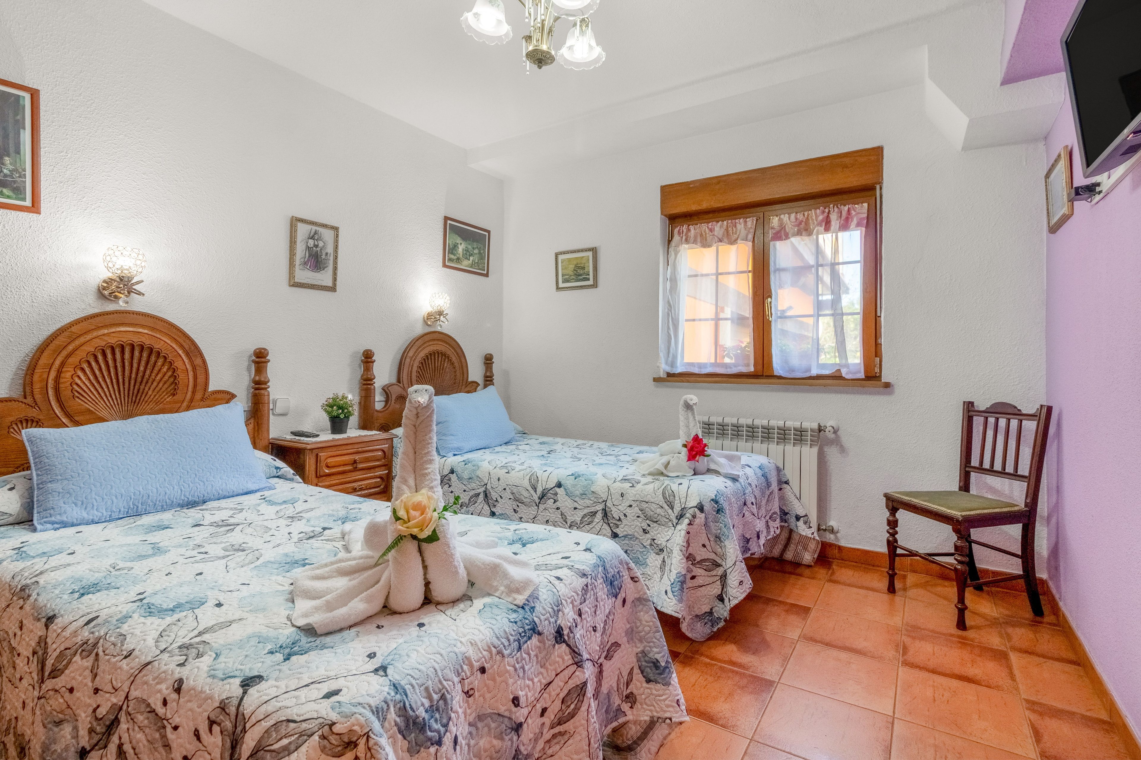 A bedroom with two beds, a wooden headboard, a window with curtains, a radiator, and a chair.
