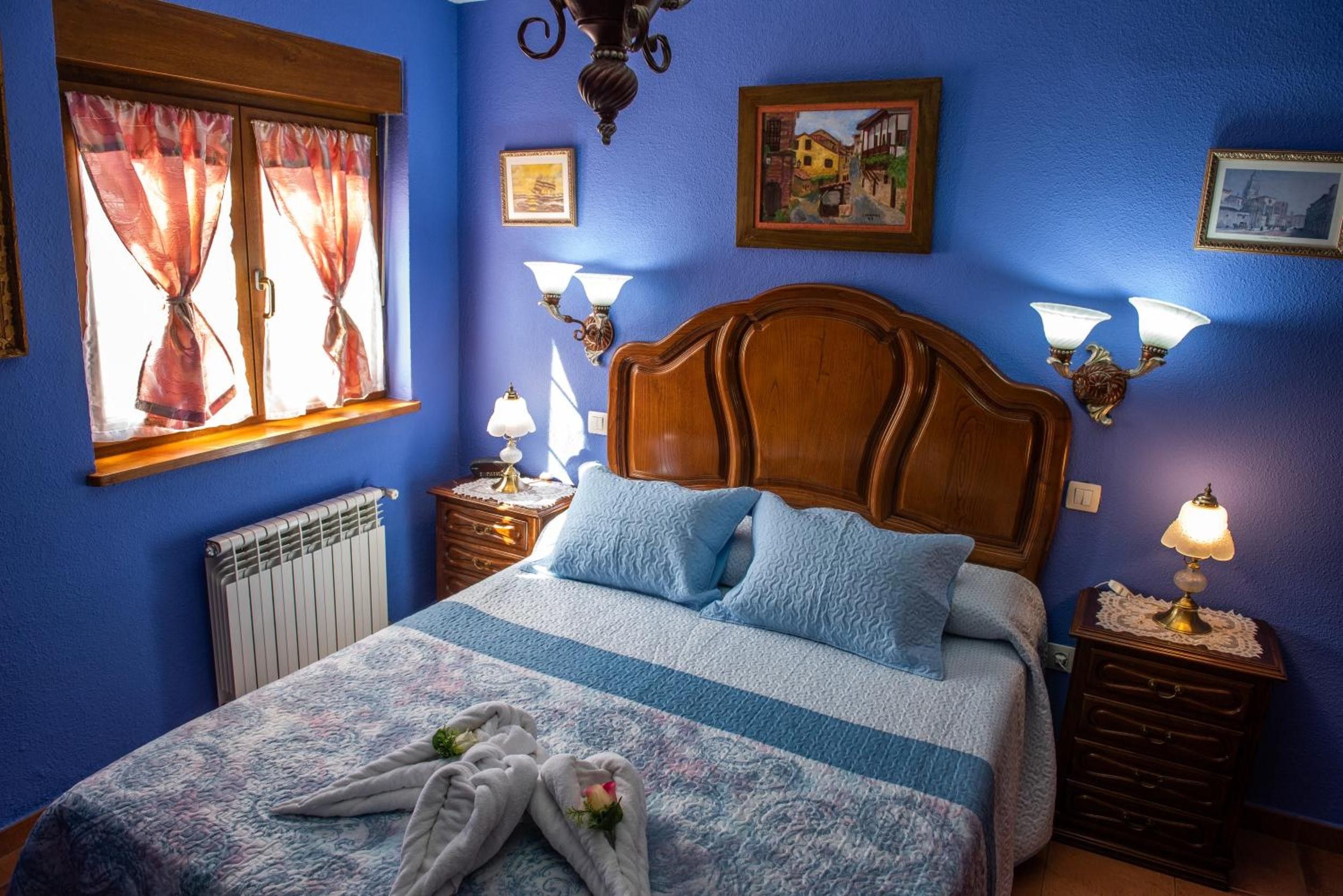 Un dormitorio con cabecera de madera, cama con ropa de cama azul y blanca, dos mesitas de noche con lámparas, una ventana con cortinas y un radiador.