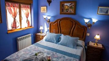 Un dormitorio con cabecera de madera, cama con ropa de cama azul y blanca, dos mesitas de noche con lámparas, una ventana con cortinas y un radiador.
